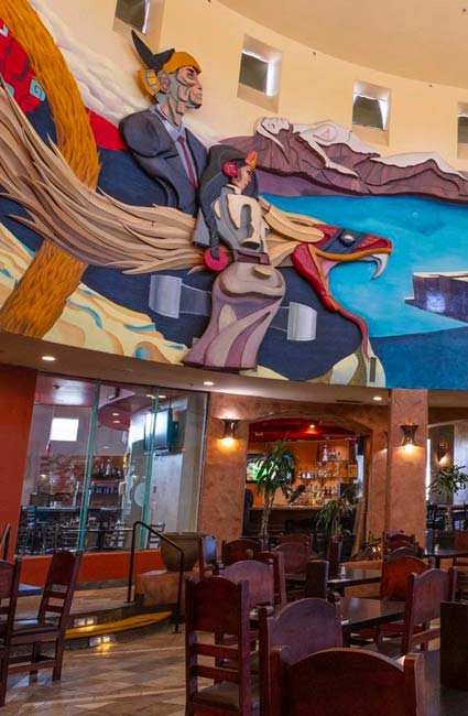Colorful mural with figures on a bird above burritos and tables at a Mexican restaurant.