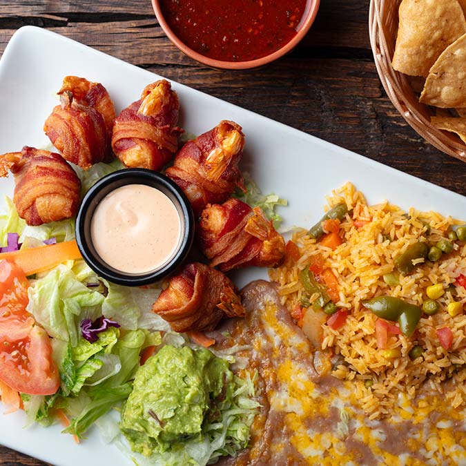 Plate with bacon-wrapped shrimp, salad, guacamole, and classic Mexican restaurant sides and sauces.