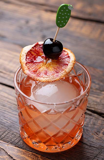 A cocktail from Escondido with a large ice sphere, blood orange slice, and cherry on a stick.