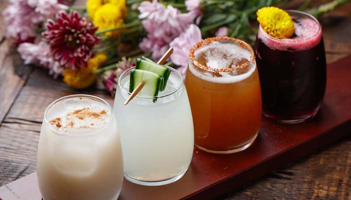 Four colorful cocktails at a Mexican restaurant, with flowers and tacos on the wooden table.