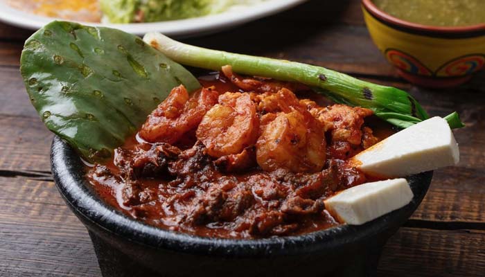 Spicy Mexican food stew with shrimp, beef, cactus leaf, onions, and cheese in a black bowl.