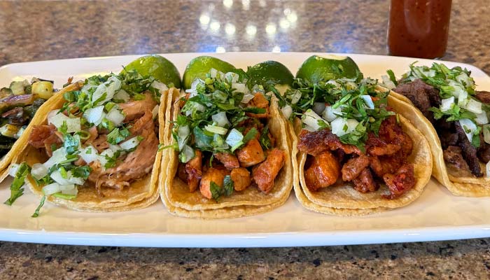 Four tacos with assorted meats, classic Mexican food topped with onions, cilantro, and fresh limes.
